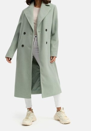 Femme portant un long manteau vert clair à double boutonnage avec un grand col, un pantalon blanc, des baskets beiges et un haut rose clair.