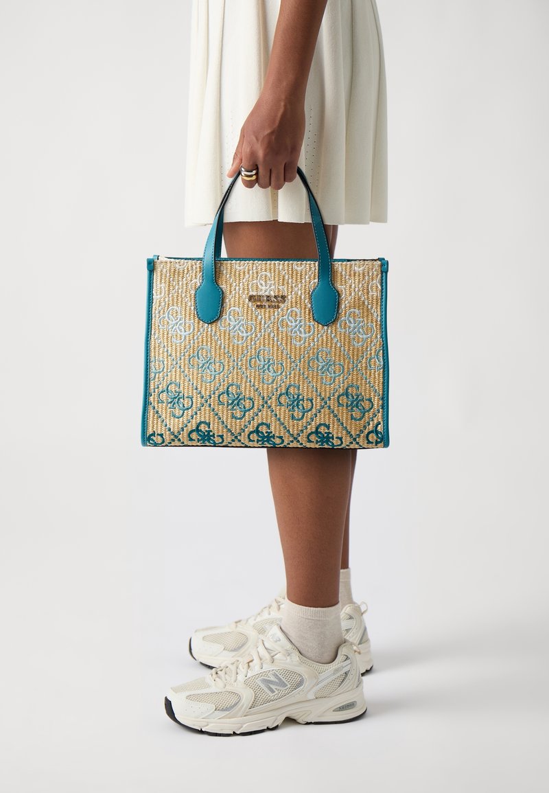Personne portant une jupe blanche et des baskets tenant un sac fourre-tout à motifs beige et bleu sarcelle par ses poignées sur le côté.