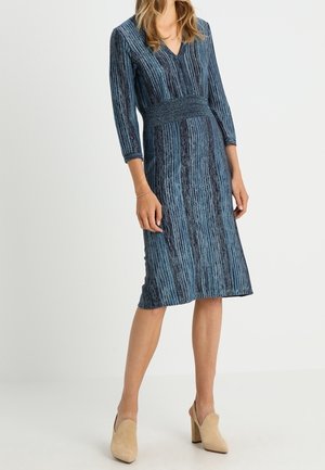 Femme portant une robe bleue et noire à rayures verticales jusqu'aux genoux, manches trois-quarts et chaussures à talons beiges.