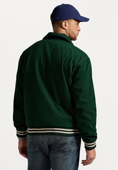Mand iført en mørkegrøn jakke med stribede manchetter og kant, blå jeans og en marineblå baseballcap, som står sidelangs mod en hvid baggrund.