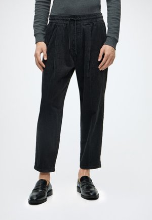 Persoon draagt zwarte, gestructureerde broek met trekkoord, donker top met lange mouwen en zwarte leren instapschoenen, staand op een witte achtergrond.