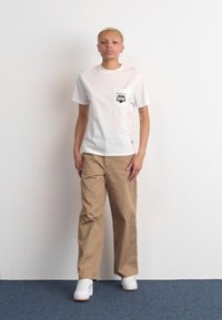 T-shirt bianco a maniche corte con taschino sul petto e grafica nera, abbinato a pantaloni larghi color beige e sneakers bianche. Tessuto in cotone morbido.