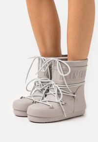 Bottes en caoutchouc grises avec une tige haute, présentant une surface supérieure texturée, des lacets décoratifs et un logo embossé sur la tige.