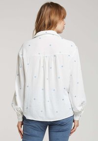 Blouse blanche avec un petit broderie florale bleue, manches longues avec des poignets boutonnés, et une coupe décontractée. Tissu texturé avec un léger éclat.