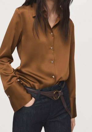 Kvinde iført en brun satin-knap-op-skjorte, stoppet ned i mørke jeans med et mørkebrunt bælte med en rund messing spænde.