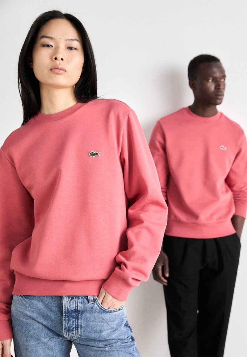 Sudaderas de cuello redondo rosa, hechas de algodón, con un pequeño logo bordado en verde. El modelo a la izquierda lleva un pantalón vaquero azul claro.