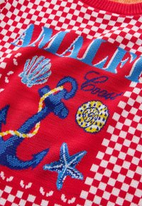 Suéter de punto rojo con un patrón a cuadros, gráficos marinos en azul, que incluyen un ancla, una concha, una estrella de mar y diseños circulares.