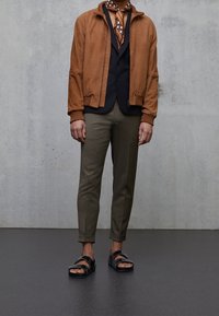 Hnědá semišová bomber bunda, černá vesta, vzorovaný šátek, olivové tapered kalhoty a černé sandály. Hladké textury, uvolněný střih, vrstvený vzhled.
