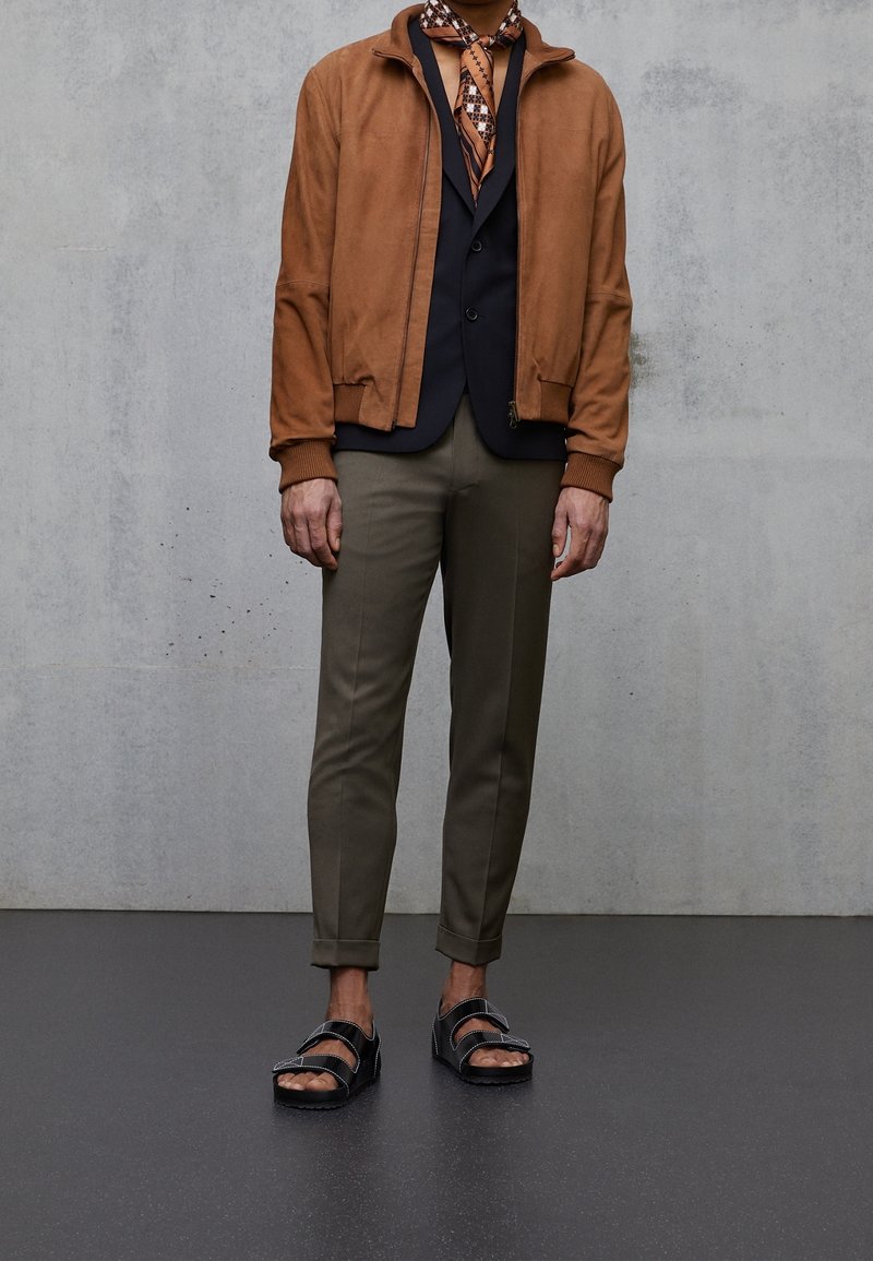Hnědá semišová bomber bunda, černá vesta, vzorovaný šátek, olivové tapered kalhoty a černé sandály. Hladké textury, uvolněný střih, vrstvený vzhled.