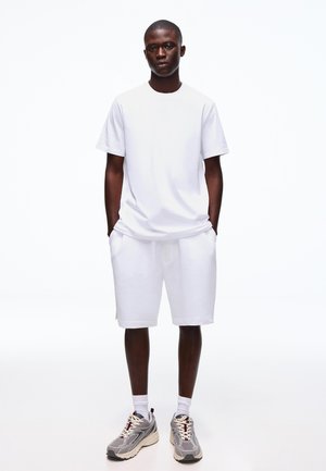Fiatal férfi zsebre tett kézzel, fehér pólót, fehér rövidnadrágot, fehér zoknit és szürke sportcipőt viselve, egyszínű fehér háttér előtt áll.