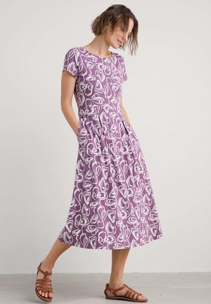 Purple dress with a white abstract pattern, short sleeves, waist pockets, and a flowing silhouette, paired with brown strappy sandals.