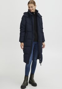 Mørkeblå dunjakke med hette, vertikalt quiltet tekstur, glidelås og to sidelommer. Kombinert med mørkejeans og kraftige svarte støvler.