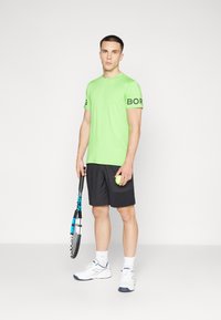 Lime zöld sport póló fekete "BORG" felirattal az ujjon, sötét rövidnadrággal kombinálva, teniszütőt és labdát tartva. Fehér cipő.