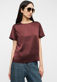 Μαρον σατέν τοπ με κοντά μανίκια και απαλή υφή, στρογγυλή λαιμόκοψη, άνετη εφαρμογή, σε συνδυασμό με ανοιχτόχρωμα τζιν, μοντέλο που φορά γυαλιά ηλίου.