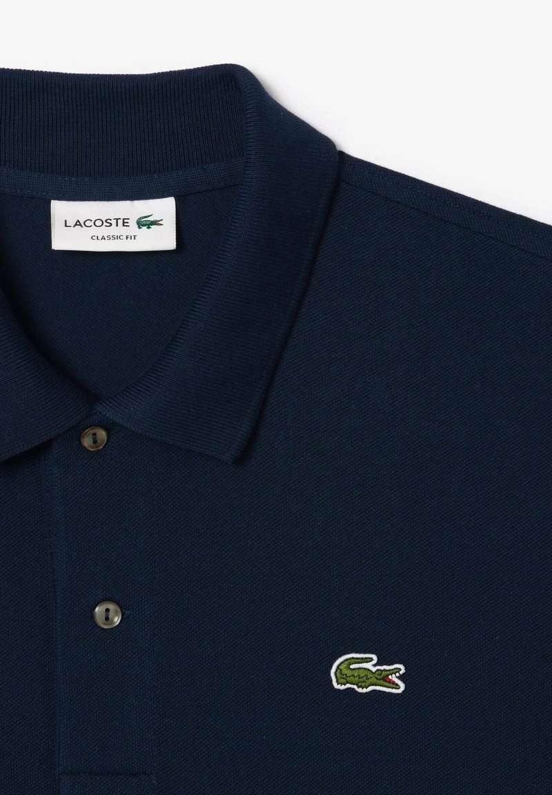 Polo Lacoste bleu marine en coton texturé, avec une patte de boutonnage à trois boutons et un logo crocodile vert sur la poitrine.
