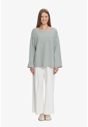 Młoda kobieta z długimi, prostymi włosami, ubrana w luźną, w paski bluzkę z długim rękawem, szerokie białe spodnie i beżowe buty wsuwane, uśmiechająca się do kamery.