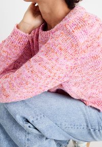 Růžový svetr s texturovaným pleteným vzorem, který obsahuje oranžové skvrnky. Kombinován se světlými modrými džínami, vytváří neformální vrstvený vzhled.