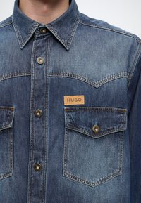 Κοντινή λήψη ενός μπλε denim πουκάμισου με μεταλλικά κουμπιά και εμπρόσθιες τσέπες, μία από τις οποίες έχει μια μικρή καφέ ετικέτα με τη λέξη HUGO.