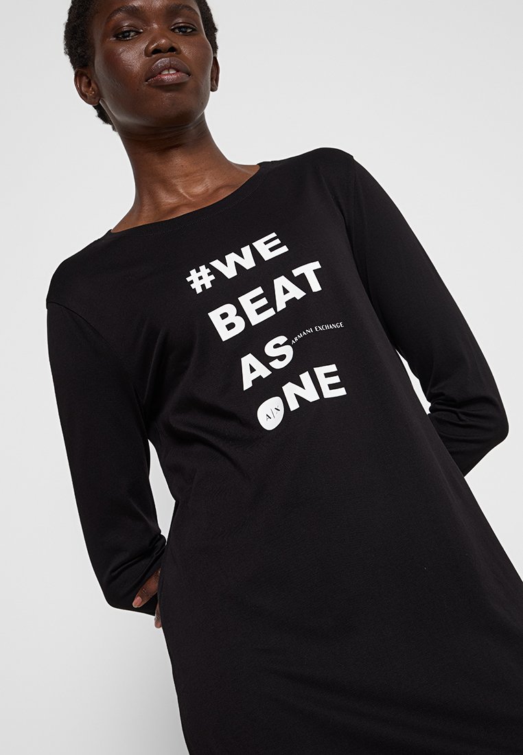 Donna che indossa una maglietta a maniche lunghe nera con il testo bianco "#WE BEAT AS ONE" e il logo Armani Exchange sul davanti.
