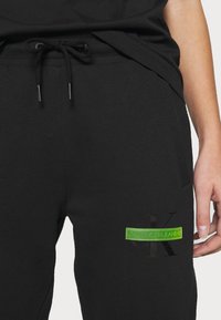 Pantalones jogger de algodón negros con cintura elástica y cordón, con una etiqueta verde que dice "Calvin Klein Jeans" y sutiles detalles de marca.
