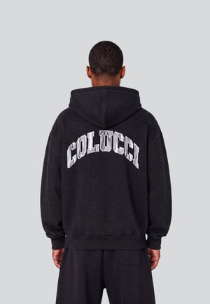 Person mit kurzen Haaren, die einen schwarzen Hoodie mit "COLUCCI" in großen weißen Buchstaben auf dem Rücken trägt, steht vor einem schlichten grauen Hintergrund.