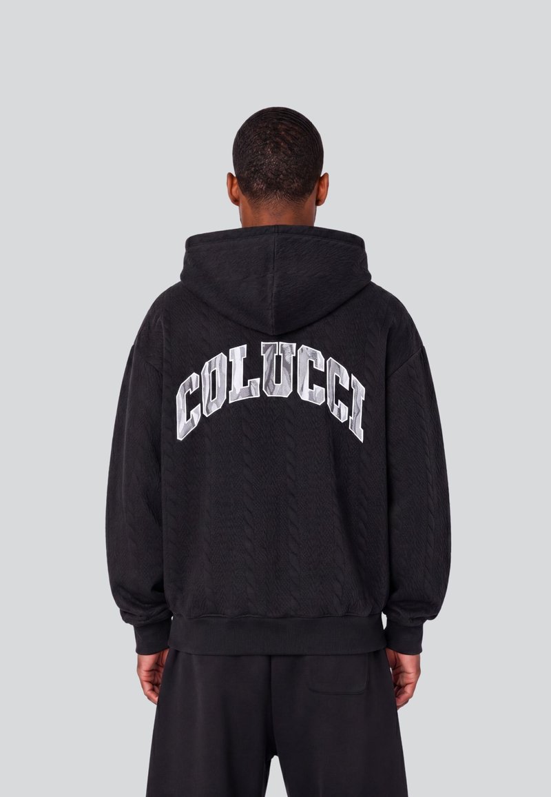 Personne aux cheveux courts portant un sweat à capuche noir avec "COLUCCI" en grandes lettres blanches dans le dos, se tenant devant un fond gris uni.