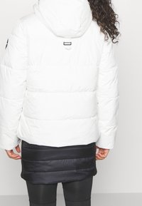 Puffig vit vinterjacka med huva, med horisontella sömmar och en svart nederdel. Designad med en ögla bak för upphängning.