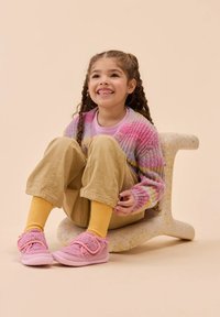 Des chaussures roses duveteuses avec une sangle en velcro, des chaussettes ribbed jaunes, un pantalon beige et un cardigan en tricot multicolore, assis sur un tabouret moucheté.