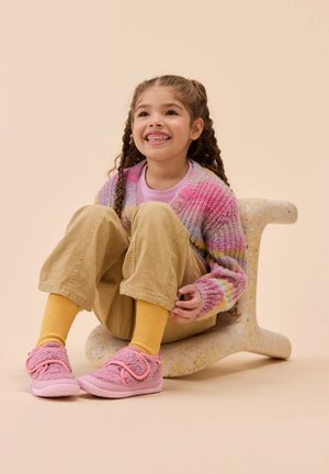 Pink fuzzy shoes with a velcro strap, yellow ribbed socks, beige pants, and a multicolored knit cardigan, seated on a speckled stool.