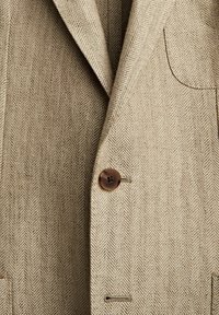 Brązowa marynarka w jodełkę z jednym brązowym guzikiem, klapą z wycięciem oraz boczną kieszenią wykonaną z fakturowanej tkaniny.