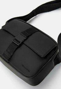 Borsa a tracolla nera realizzata in materiale liscio, con pattina pieghevole e chiusura a fibbia, tracolla regolabile e logo del brand in rilievo.