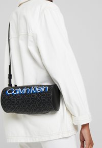 Borsa rotonda nera con motivo ripetuto "CK", con logo blu "Calvin Klein" e tracolla regolabile. Texture liscia.