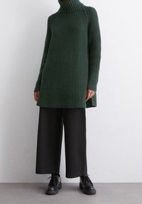 Dunkelgrüner, strukturierter Rollkragenpullover mit lockerer Passform und seitlichen Schlitzen. Kombiniert mit weiten schwarzen Hosen und schwarzen Schnürschuhen.