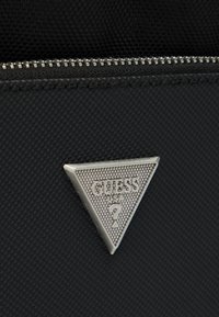 Μαύρο υφασμάτινο τσαντάκι με ασημένιο φερμουάρ και τριγωνική μεταλλική πινακίδα λογότυπου με την επιγραφή "GUESS? USA" στο κέντρο.