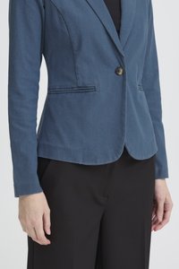 Naine kannab keha vormivat sinist jopet ühe nööbiga ja musti pükse, seisab ühevärvilise tausta ees.