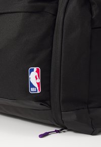 Zwarte nylon rugzak met een meerkleurige NBA-logo patch. De rits heeft een paarse trekker en een gestructureerde afwerking.