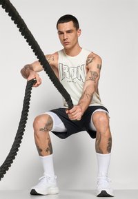 Athletic man in a sleeveless cream tank top with "IRON" print, black shorts, and white shoes, gripping a black battle rope. Visible tattoos.