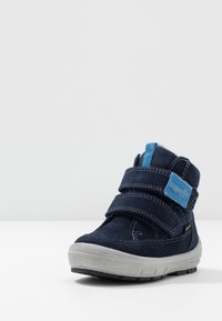 Botín de ante azul marino con dos correas ajustables de Velcro, suela de goma gris y acento de marca azul. Superficie texturizada y detalles de costura.