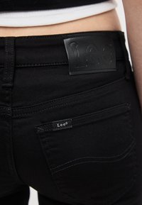 Κοντινό πλάνο της πίσω τσέπης και της ζώνης μέσης μαύρων τζιν Lee με ετικέτες με το λογότυπο Lee σε άτομο που φοράει λευκό τοπ.