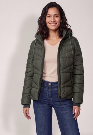 Femme souriante aux cheveux foncés porte une veste matelassée vert olive, une chemise beige clair et un jean bleu sur un fond clair uni.