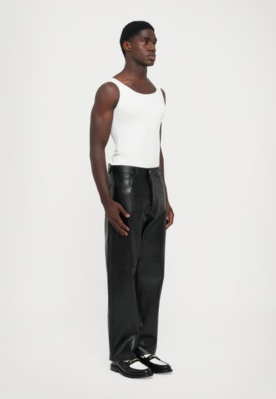 Homme debout portant un débardeur blanc sans manches, un pantalon en cuir large noir et des mocassins noir et blanc sur un fond uni.
