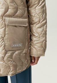 Beige prošívaná bunda s kapsou, vyznačující se hladkou texturou a vlnkovým vzorem stehu. Na kapse je viditelná nášivka s logem.