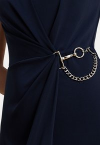 Jūrinė suknelė, pasižyminti susiraukiamu audiniu ir sidabro grandine su užsegimu, sukurianti tekstūrą ir vizualinį interesą.