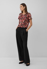 Kurzärmlige Bluse in Pink und Rot mit Muster, kombiniert mit schwarzen plissierten Hosen; das Model steht mit Händen in den Taschen und trägt schwarze Ballerinas.