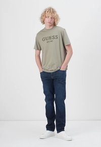 Maslinasto zelena majica s natpisom "GUESS EST.1981" u crnoj boji, uparena s tamnoplavim trapericama i bijelim tenisicama. Model stoji naslonjen na neutralnu pozadinu.