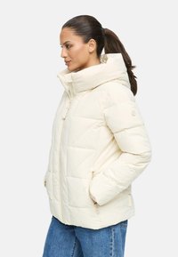 Veste matelassée beige clair avec un col montant, un design en biais, une fermeture éclair à l'avant et des poches latérales. Tissu doux avec une texture lisse.