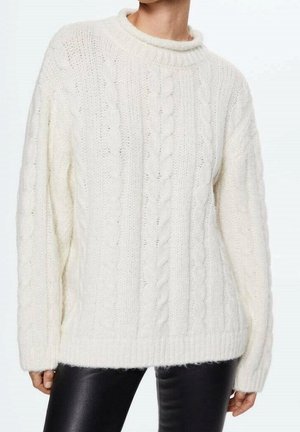 Femme portant un pull blanc à torsades avec manches longues et un pantalon en cuir noir, se tenant devant un fond blanc uni.