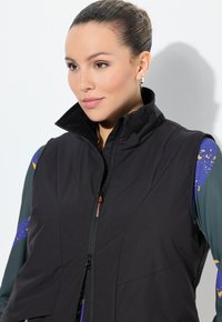 Schwarze ärmellose Weste mit hohem Kragen, ausgestattet mit einem Frontreißverschluss und flexiblem Material, kombiniert mit einem bunten gemusterten Langarmshirt.