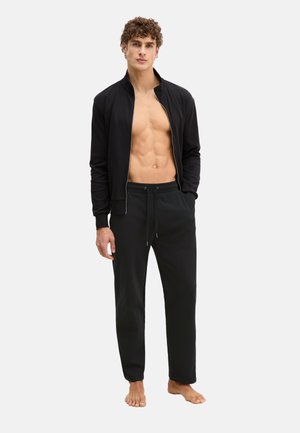 Junger Mann steht barfuß da, trägt eine schwarze Jacke mit Reißverschluss, die offen ist und die nackte Brust sowie schwarze Hosen mit Kordelzug vor weißem Hintergrund zeigt.