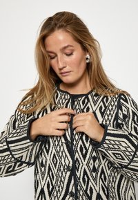 Czarna i kremowa kurtka w geometryczne wzory, z zaokrąglonym kołnierzem i zapięciem na guziki; strukturalna tkanina z kontrastowym wykończeniem.